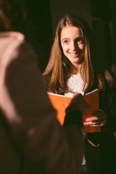 Schülerin hält ein Skript in den Händen und lächelt während einer Szene in einer Schultheaterprobe – erste Erfahrungen auf dem Weg zur Schauspielausbildung.
